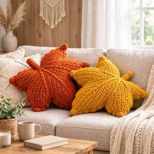 May include: Two chunky knit throw pillows shaped like maple leaves, one in a vibrant burnt orange and the other in a mustard yellow, resting on a light-colored sofa alongside a white textured throw blanket and a small potted plant.