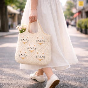 Può includere: Borsa tote all'uncinetto color crema con facce di gatto ricamate e colletti gialli. La borsa ha un motivo a rombi e manici robusti. La borsa è piena di fiori rosa ed è portata da una persona che indossa un vestito e scarpe bianche.