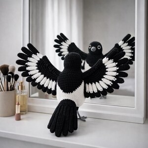 May include: A crocheted magpie with black and white wings spread wide, reflected in a mirror. The bird's body is black and white. The bird is on a white surface, with makeup and perfume in the background. The bird's wingspan is approximately 30 cm.