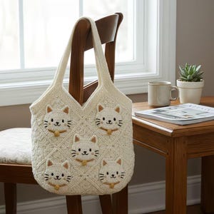 May include: Cream-colored crochet tote bag featuring five embroidered cat faces. The bag has a diamond pattern and a sturdy handle. The cat faces have brown ears, whiskers, and a small gold-colored collar. The bag is hanging on a wooden chair.
