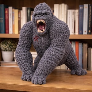 May include: A handmade, gray crochet gorilla with an open mouth, revealing teeth and a pink interior. The gorilla has a red stitched scar on its chest. The toy is posed on all fours, with a bookshelf in the background.