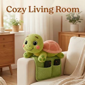 Op de afbeelding: Een groene schildpadknuffel met een roze schild zit op een groene organizer met vakken, met twee zwarte afstandsbedieningen. De woorden "Cozy Living Room" staan boven de schildpad. De schildpad staat op een witte bank.