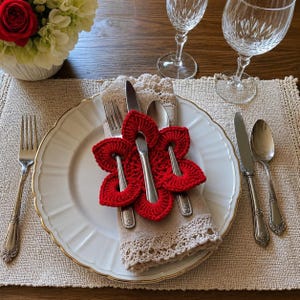 May include: A bright red crocheted flower napkin ring holds a silver fork, knife, and spoon against a beige linen napkin with lace trim. The set rests on a white plate with a gold rim, placed on a textured beige placemat with silverware nearby.