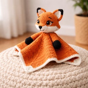 K&ouml;nnte beinhalten: Ein handgefertigtes Baby-Kuscheltuch mit einem geh&auml;kelten Fuchs. Der Fuchs hat orangefarbenes Fell mit wei&szlig;en und schwarzen Akzenten. Der Deckenbereich ist orange mit wei&szlig;em Rand. Der Fuchs liegt auf einer cremefarbenen, geh&auml;kelten Oberfl&auml;che.