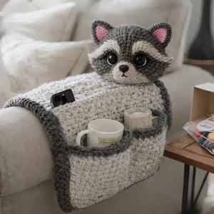 Pode incluir: Apoio de braço de sofá de crochê feito à mão com cabeça de guaxinim e bolsos. O apoio de braço cinza e branco tem dois porta-copos e um bolso para um telefone. O guaxinim tem pelo cinza e branco com orelhas cor-de-rosa.