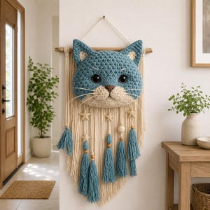 May include: A blue and beige crochet cat wall hanging. The cat face is centered on a cream-colored macrame design with blue tassels, wooden beads, and star accents. The cat has large black eyes and a beige nose and mouth.