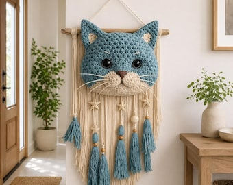 May include: A blue and beige crochet cat wall hanging. The cat face is centered on a cream-colored macrame design with blue tassels, wooden beads, and star accents. The cat has large black eyes and a beige nose and mouth.