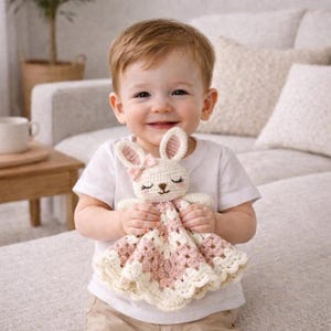 May include: A cream, pink, and white crocheted bunny comforter. The bunny features pink inner ears, a pink bow, and embroidered facial details. The blanket has a scalloped edge and a pink and white checked pattern. A child is holding the comforter.