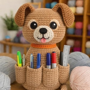 May include: A handmade, crocheted dog-shaped desk organizer in shades of brown, with a white muzzle, black eyes, and a pink tongue. The dog has pockets holding pens, markers, and a ruler. An orange collar accents the design. Yarn balls are in the background.