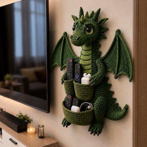 May include: A dark green crocheted dragon wall organizer featuring two integrated pockets on its belly. The dragon has large dark eyes, textured scales, and wide wings. The pockets contain multiple black remote controls, white earbuds, and a charging cable.