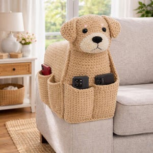 May include: A tan crochet dog-shaped couch arm organizer with multiple pockets. The dog has a cream-colored snout and black eyes. Several smartphones are placed in the pockets. The organizer is placed on the arm of a light gray couch.