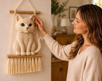 May include: A cream-colored, knitted cat wall hanging. The cat has green eyes and a long tail. It is suspended from a wooden dowel with a macrame-style fringe. The cat is sitting on a wooden shelf. The wall hanging is displayed on a white wall.