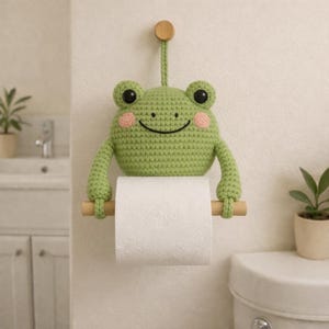 May include: A green crocheted frog toilet paper holder with a wooden dowel. The frog has black eyes and pink cheeks. The holder is hanging on a white wall in a bathroom setting.