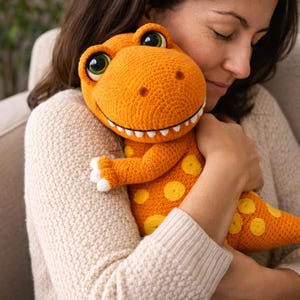 May include: An orange crocheted dinosaur stuffed animal with yellow spots and white teeth. The dinosaur has large, expressive eyes with green irises. The toy is being held close, suggesting a soft and cuddly texture.