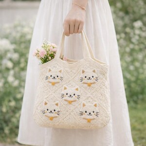 May include: Cream-colored, crocheted tote bag featuring embroidered cat faces. The bag has a diamond pattern and a small bouquet of pink flowers inside. The cat faces have black whiskers and eyes, and a yellow collar. The bag is held by a person wearing a white dress.