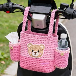 May include: Pink crocheted handlebar bag with a bear applique. The bag has two side pockets, one holding tissues and the other a water bottle. The bag is designed to attach to a scooter or bicycle handlebar.