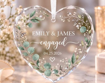 Puede incluir: Un adorno de cristal transparente en forma de corazón facetado que cuelga de una cinta blanca. El adorno presenta una corona impresa de hojas de eucalipto verdes y flores blancas, con el texto EMILY & JAMES, engaged, y la fecha 25.04.2026 en blanco.