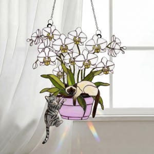 Puede incluir: Arte colgante de vidrieras con una orqu&iacute;dea en maceta con flores blancas y hojas verdes. Un gato siam&eacute;s descansa en la maceta, mientras que un gato atigrado gris trepa por el costado. La maceta es lavanda y el arte cuelga de cadenas.