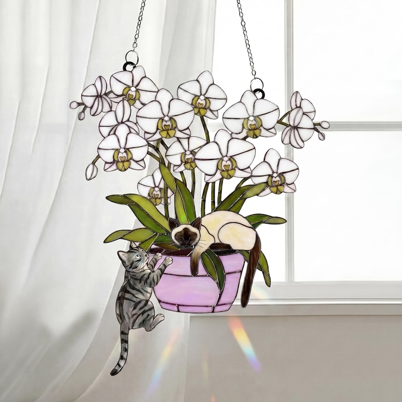 Puede incluir: Arte colgante de vidrieras con una orqu&iacute;dea en maceta con flores blancas y hojas verdes. Un gato siam&eacute;s descansa en la maceta, mientras que un gato atigrado gris trepa por el costado. La maceta es lavanda y el arte cuelga de cadenas.