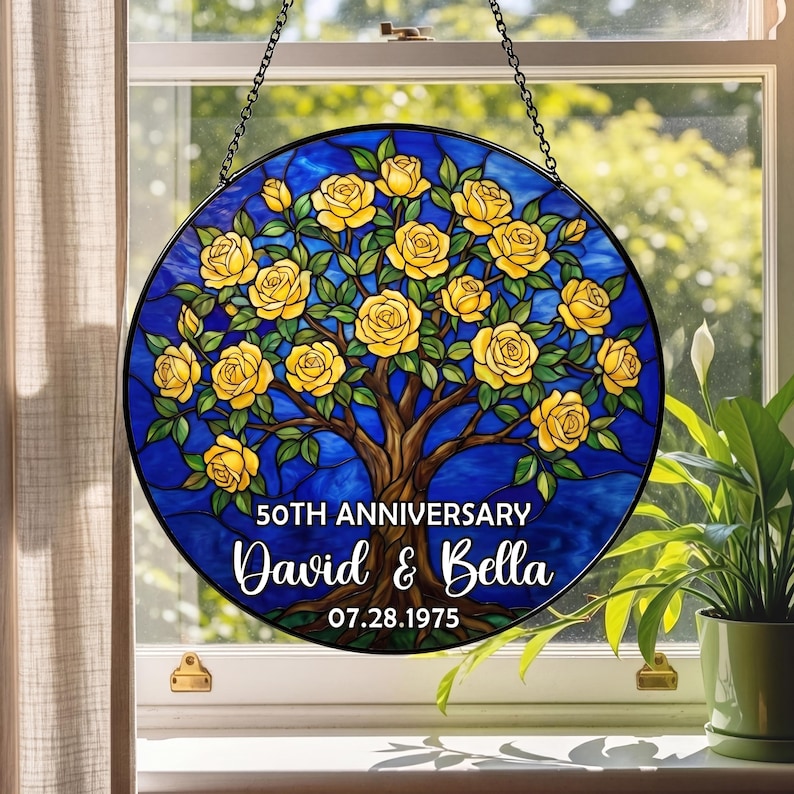 May include: Round stained glass window art with a blue background and a tree with yellow roses. The text reads "50th Anniversary" and the names "David & Bella" with the date "07.28.1975". The artwork is hanging in front of a window.