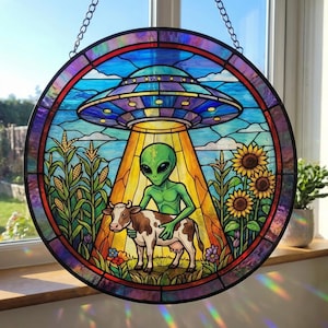 May include: Round stained glass window featuring a green alien with a cow under a UFO. The scene includes corn stalks, sunflowers, and a blue sky. The window is framed with a chain for hanging.