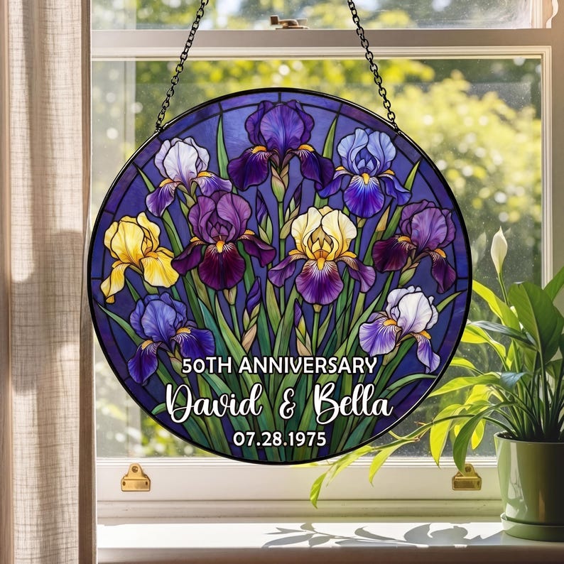 May include: A round stained glass window featuring a floral design of irises in shades of purple, yellow, and white. The text "50th Anniversary David & Bella 07.28.1975" is displayed on the bottom. The window is hanging in front of a window.