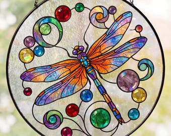 Puede incluir: Una vidriera redonda con un diseño de libélula colorida. La libélula tiene alas naranjas, azules y moradas. La ventana está decorada con cuentas y espirales de vidrio de colores, y está enmarcada con un borde de metal negro.