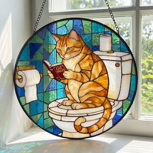 May include: Round stained glass window art featuring an orange tabby cat reading a book titled "CAT TALES" while seated on a toilet. The background is a mosaic of blue and green glass. A roll of toilet paper is on the left.