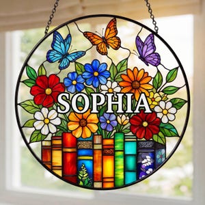 May include: A round, stained-glass window with a black metal frame, featuring colorful flowers, butterflies, and the name "SOPHIA" in white. The design includes a row of books at the bottom, with a chain for hanging.