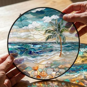 Puede incluir: Obra de arte redonda de vidrieras que representa una escena de playa. La obra presenta una palmera, olas del oc&eacute;ano, nubes blancas y conchas marinas. El vidrio es en tonos de azul, verde y beige, con un marco de metal negro. La pieza est&aacute; sostenida por dos manos.