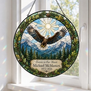 May include: A circular stained glass suncatcher featuring a bald eagle in flight over a mountain range and pine forest. The border is decorated with oak leaves and pinecones. Text reads: Forever in Our Hearts, Michael McMasters, 1972-2026.