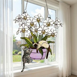 Puede incluir: Colgante de ventana de vidrieras con una planta de orqu&iacute;deas blancas en una maceta morada, con dos gatos. Un gato est&aacute; trepando por la maceta, mientras que el otro est&aacute; descansando. El arte de la ventana est&aacute; suspendido por cadenas.