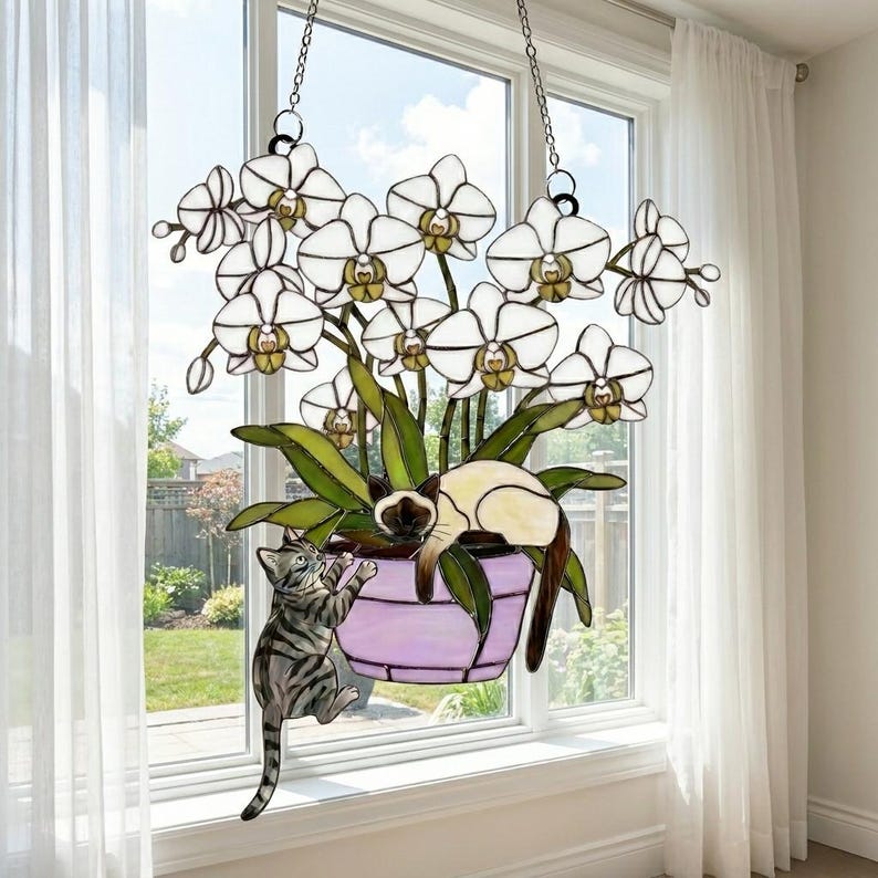 Puede incluir: Colgante de ventana de vidrieras con una planta de orqu&iacute;deas blancas en una maceta morada, con dos gatos. Un gato est&aacute; trepando por la maceta, mientras que el otro est&aacute; descansando. El arte de la ventana est&aacute; suspendido por cadenas.