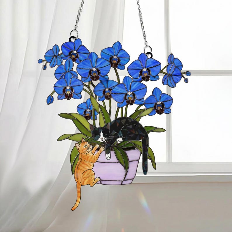 Puede incluir: Arte colgante de vidrieras con una vibrante planta de orqu&iacute;deas azules en una maceta lavanda, con un gato blanco y negro descansando sobre las hojas y un gato atigrado naranja trepando por la maceta. Suspendido por cadenas plateadas.