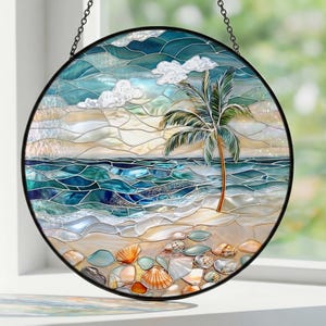 Puede incluir: Vidriera redonda que representa una escena de playa con una palmera, el oc&eacute;ano y nubes. La obra de arte tiene un marco de metal negro y una cadena para colgar. La playa est&aacute; adornada con conchas marinas y guijarros.