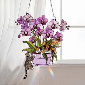 Puede incluir: Un macetero colgante de vidrieras con orqu&iacute;deas moradas y gatos. Los gatos est&aacute;n representados trepando y descansando. El macetero est&aacute; suspendido por cadenas, creando una pieza decorativa. Las orqu&iacute;deas est&aacute;n en una maceta lila.