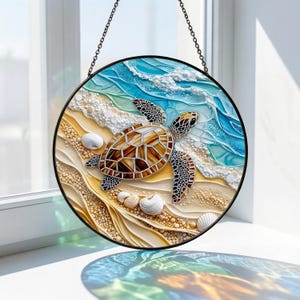May include: Round stained glass art piece featuring a sea turtle swimming in the ocean. The turtle has a brown and tan shell, with a black border. The background depicts a beach scene with blue waves, sand, and seashells. The piece is hanging by a black chain.