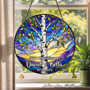 May include: A round stained glass window hanging with a tree design. The tree has a white trunk and branches with green and yellow leaves. The background is blue, purple, and yellow. Text reads "50th Anniversary David & Bella 07.28.1975".