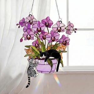 Puede incluir: Un adorno colgante de vidrieras con una planta de orqu&iacute;deas moradas en una maceta lavanda, con varios gatos. Un gato blanco y negro descansa sobre las hojas, mientras que otros se asoman. Un gato atigrado gris trepa por la maceta.