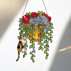 Puede incluir: Arte colgante de vidrieras que representa a un gato durmiendo en una maceta con flores rojas y enredaderas verdes. Otro gato est&aacute; trepando por la maceta. La maceta es naranja y est&aacute; suspendida por una cadena.