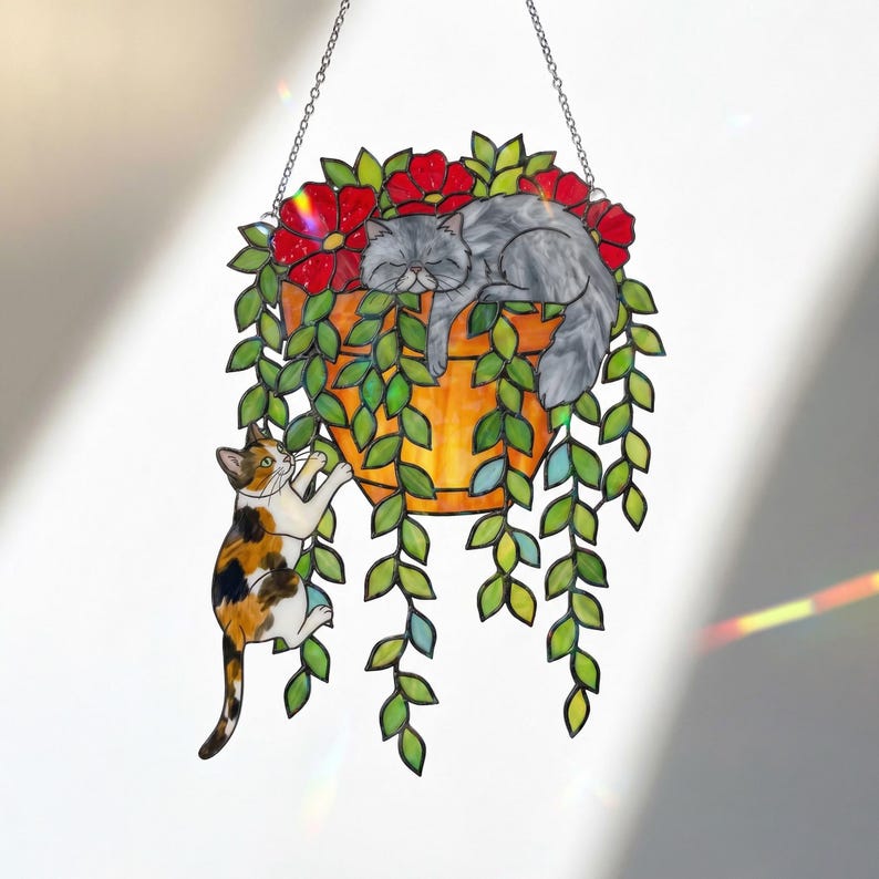 Puede incluir: Arte colgante de vidrieras que representa a un gato durmiendo en una maceta con flores rojas y enredaderas verdes. Otro gato est&aacute; trepando por la maceta. La maceta es naranja y est&aacute; suspendida por una cadena.