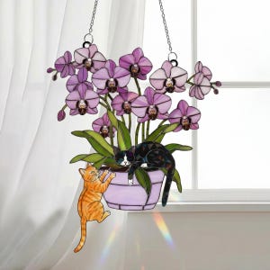 Puede incluir: Un adorno colgante de vidrieras con una planta de orqu&iacute;deas moradas en una maceta lavanda. Se representan dos gatos, uno naranja y otro blanco y negro, interactuando con la planta. El adorno cuelga de dos cadenas plateadas.