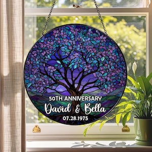 May include: Round stained glass panel featuring a tree design in shades of purple, blue, and green. The panel includes the text "50TH ANNIVERSARY" above the names "David & Bella" and the date "07.28.1975". The panel is hanging in front of a window.