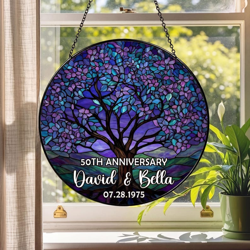 May include: Round stained glass panel featuring a tree design in shades of purple, blue, and green. The panel includes the text "50TH ANNIVERSARY" above the names "David & Bella" and the date "07.28.1975". The panel is hanging in front of a window.