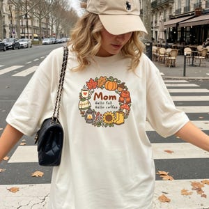 Puede incluir: Una camiseta color crema con un gráfico circular de temática otoñal que incluye una calabaza, hojas, girasol, linterna, suéter, taza y botas. El texto dice Mom, Hello fall, Hello coffee. Se lleva un bolso de hombro negro acolchado.