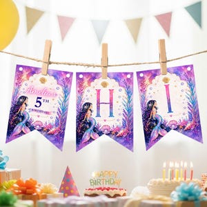 Puede incluir: Una pancarta de cumplea&ntilde;os con tem&aacute;tica de sirenas con el texto "Amelia's 5th Birthday" en tres banderines. La pancarta est&aacute; decorada con sirenas, conchas marinas y una combinaci&oacute;n de colores p&uacute;rpura y azul. La pancarta est&aacute; sujeta con pinzas de madera.