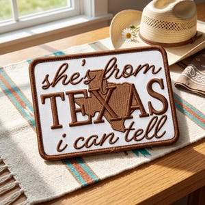 May include: A rectangular white patch with brown embroidered text that reads "she's from TEXAS i can tell". The patch has a brown border and a brown embroidered outline of the state of Texas. A straw hat and a patterned scarf are in the background.