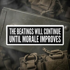 May include: A black and white rectangular sticker with the text "THE BEATINGS WILL CONTINUE UNTIL MORALE IMPROVES" in bold white letters. The sticker is set against a backdrop of military gear, including a green metal box and tactical equipment.