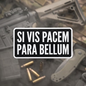May include: A black and white rectangular sticker with the Latin phrase "Si Vis Pacem Para Bellum" in bold white letters. The sticker is set against a blurred background of firearms, ammunition, and a folding knife.