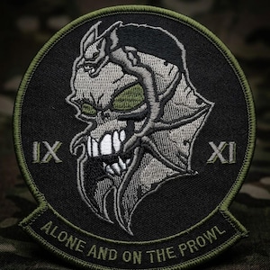 May include: Embroidered patch featuring a stylized skull with green eyes and a serpentine design. The patch is black with a green border and the text "ALONE AND ON THE PROWL". Roman numerals IX and XI are also present.